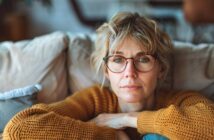 a middle-aged woman wearing glasses and a knitted orange jumper sits on her couch with a bored expression on her face