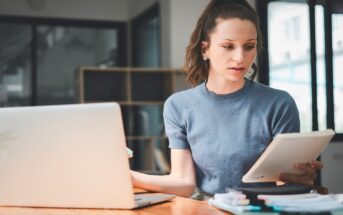 a young brunette woman wearing a blue top working from her home office. She is looking at some papers with her laptop to one side