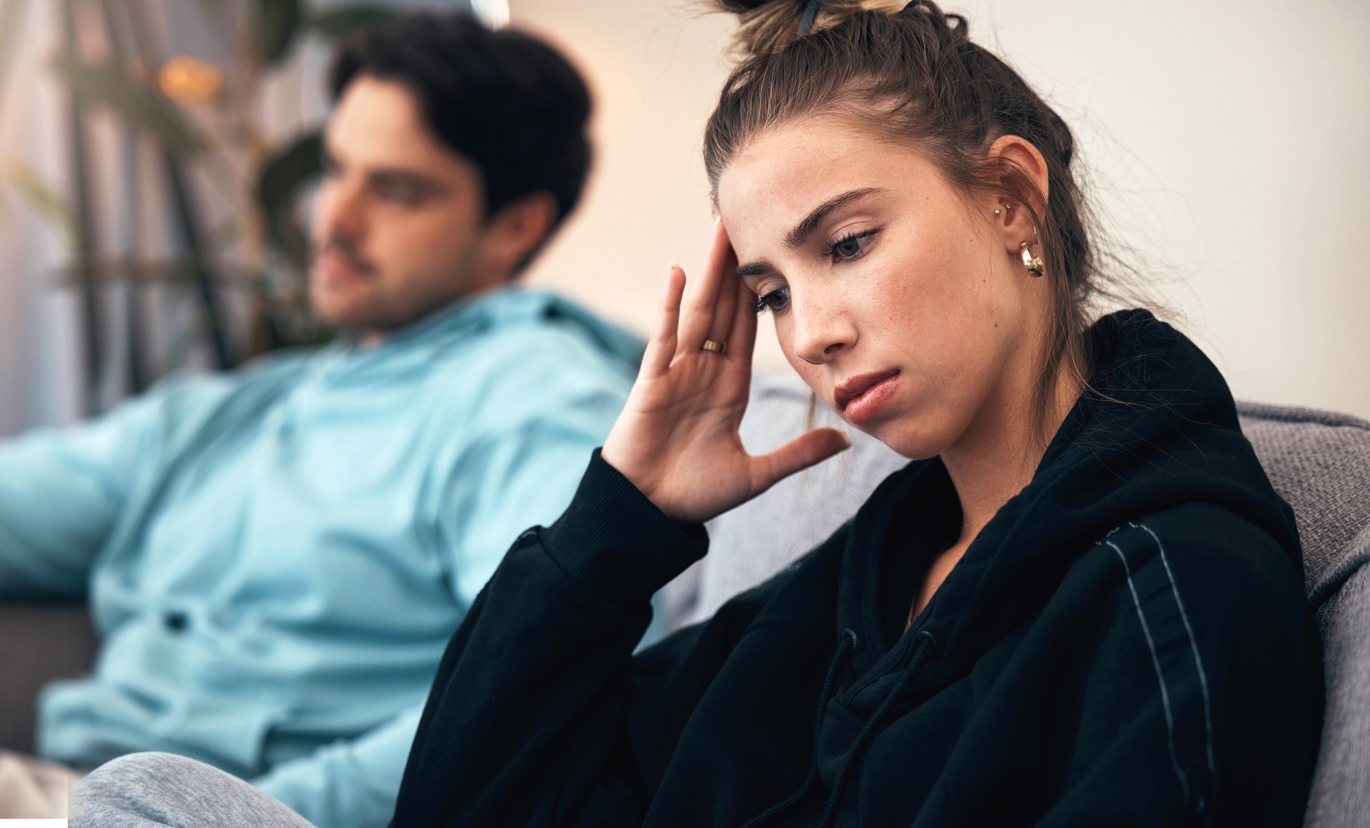 A young woman sits on a couch in the foreground, looking distressed with her hand on her forehead. A young man is sitting next to her in the background, slightly out of focus, appearing concerned. The room has muted lighting and a calm ambiance.