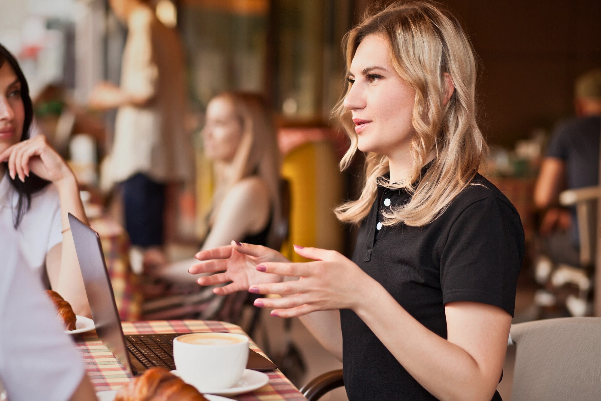 A woman with blonde hair, wearing a black shirt, sits at an outdoor café table, gesturing expressively with her hands while engaging in conversation. A laptop and a cup of coffee are on the table in front of her. Other patrons are seen in the background.
