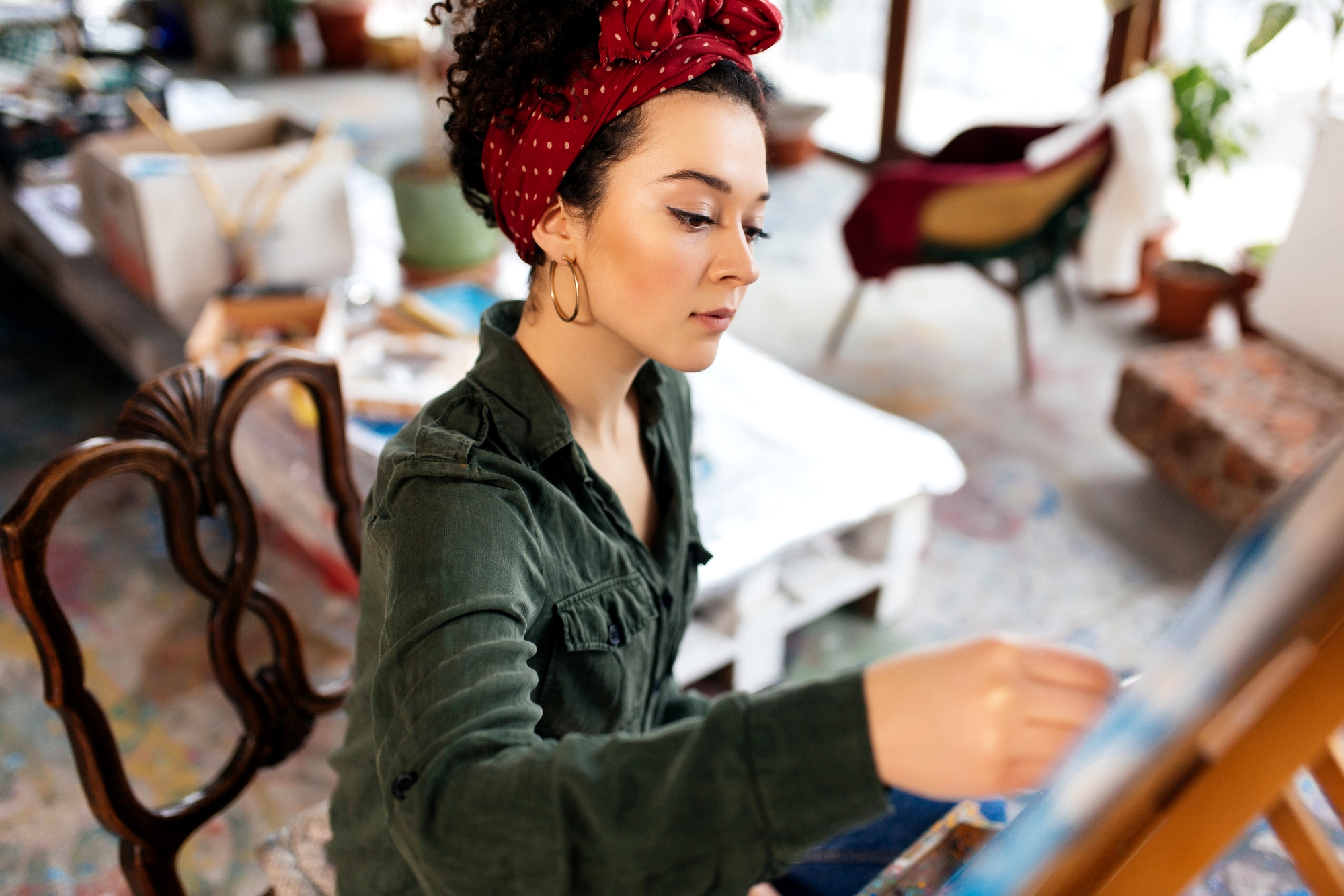 A woman with a red headband and hoop earrings is focused on painting. She is seated in a studio with various art supplies and furniture in the background. She wears a green shirt and holds a brush, working on a canvas. The environment is cozy and artistic.