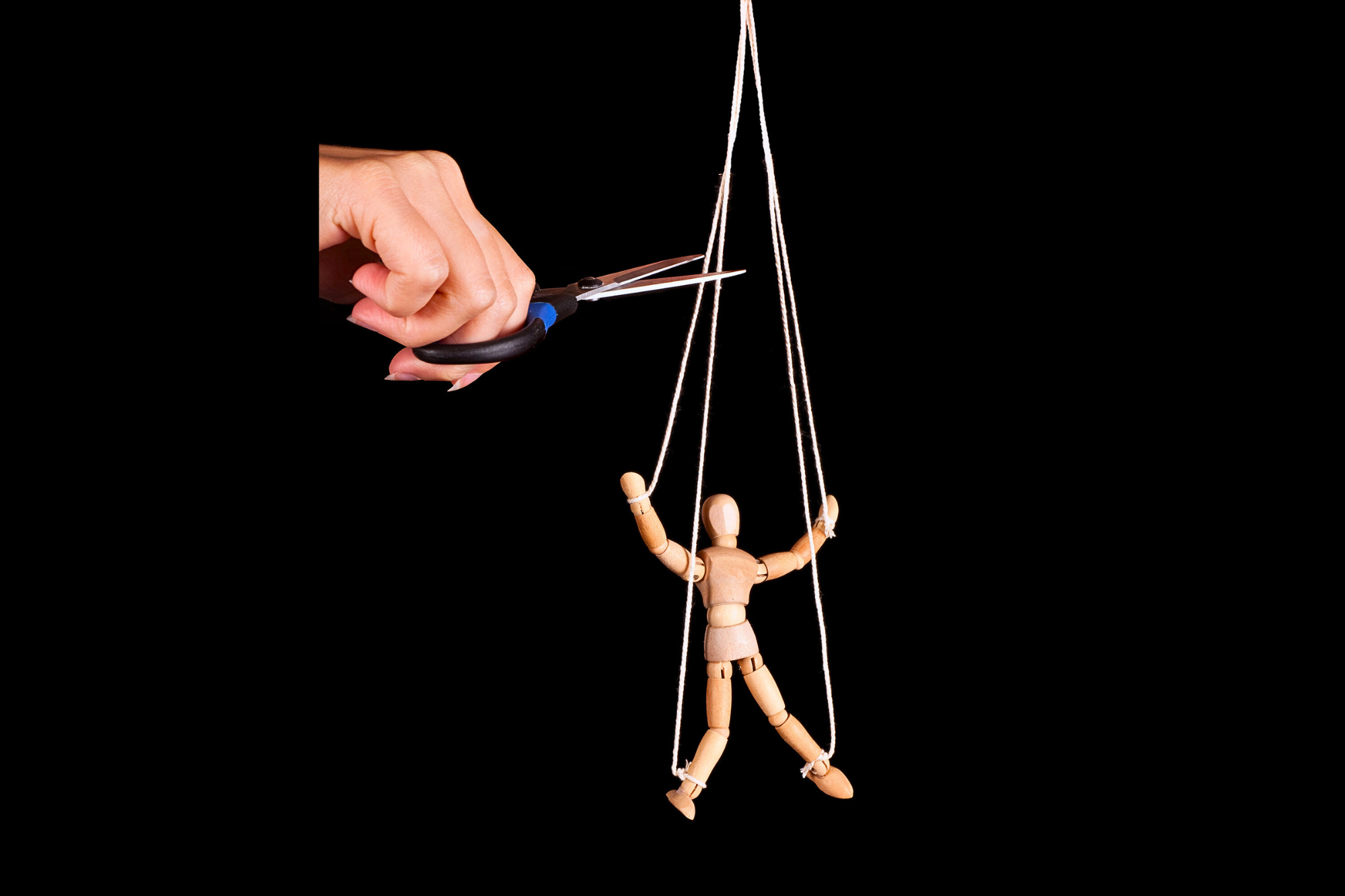 A wooden marionette puppet hangs by strings against a black background. A human hand holding a pair of scissors is positioned to cut the strings, symbolizing breaking free from control or constraints.