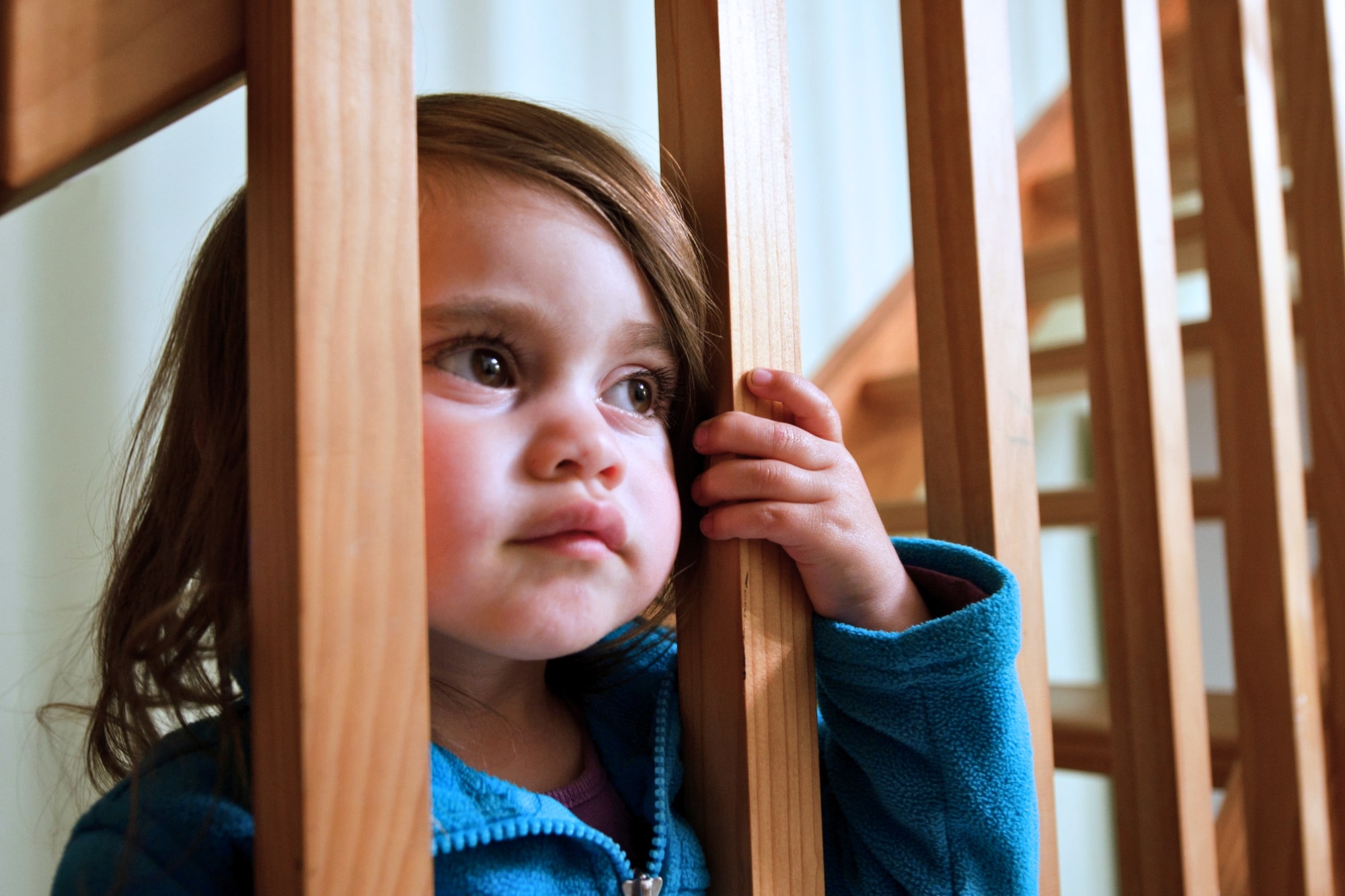 A young child with long brown hair and wearing a blue jacket looks thoughtfully through wooden stair railings, holding one rail with her hand. The background is softly lit, highlighting the wooden texture of the stairs.