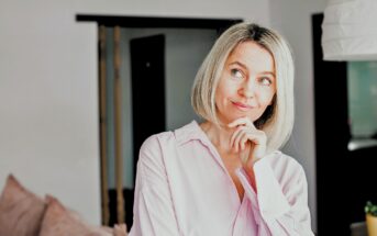 A woman with blonde hair and a light pink shirt stands indoors, resting her chin on her hand and smiling thoughtfully, with a blurred background of a modern living space.