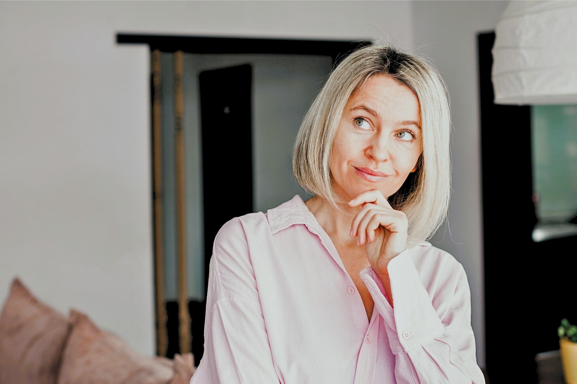 A woman with blonde hair and a light pink shirt stands indoors, resting her chin on her hand and smiling thoughtfully, with a blurred background of a modern living space.