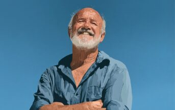 An older man with a white beard and mustache smiles confidently with arms crossed, wearing a blue shirt, standing outdoors against a clear blue sky.