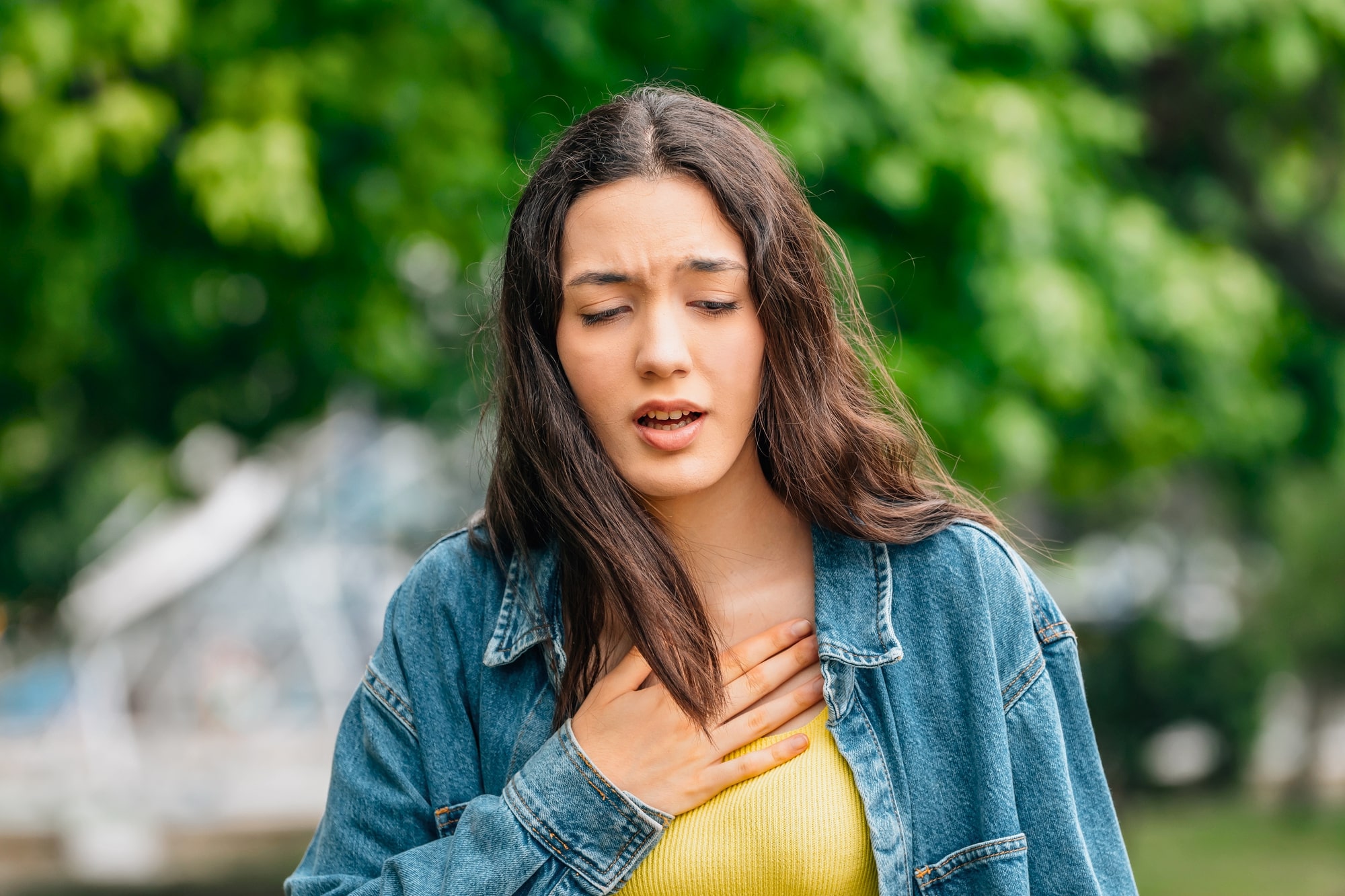 A woman with long brown hair, wearing a yellow top and a denim jacket, stands outdoors with her eyes partially closed. She looks distressed and is holding her chest with one hand. The background features lush green trees.