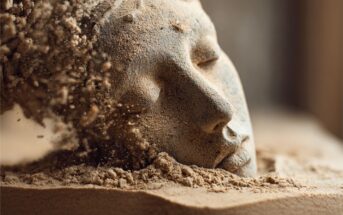 A detailed sand sculpture of a serene face is partially buried, with sand dispersing from one side, creating a sense of erosion or disintegration.