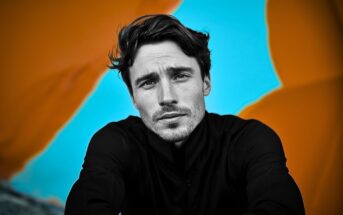 A man in a black shirt looks into the camera with a serious expression; the background is bright blue with large, blurred orange shapes.