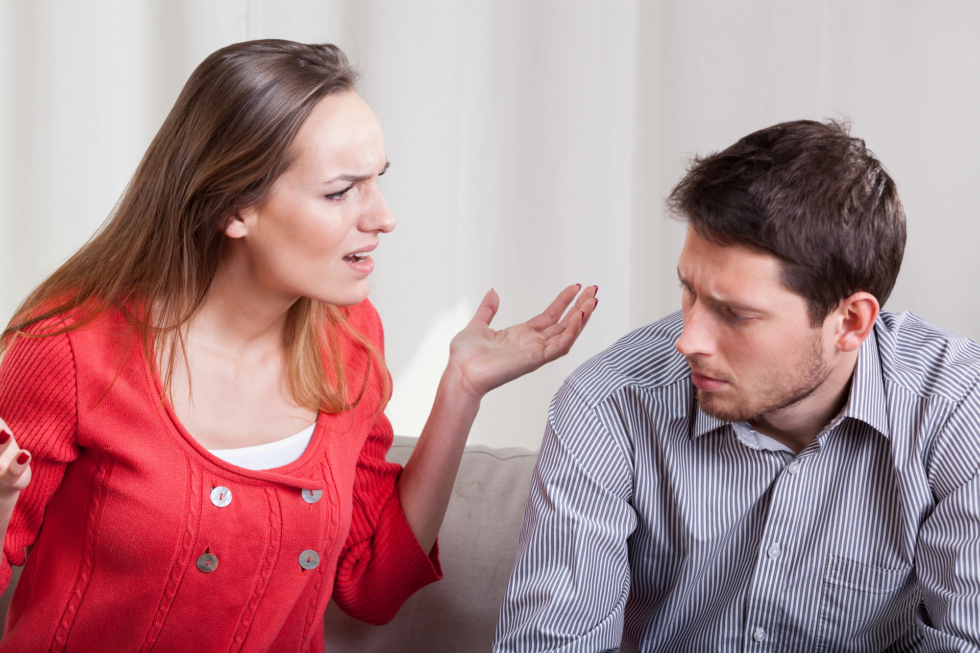 A woman wearing a red sweater with a frustrated expression and raised hands is talking to a man in a striped shirt. The man, sitting beside her, has a troubled look and is staring downward. The setting appears to be indoors with a plain background.
