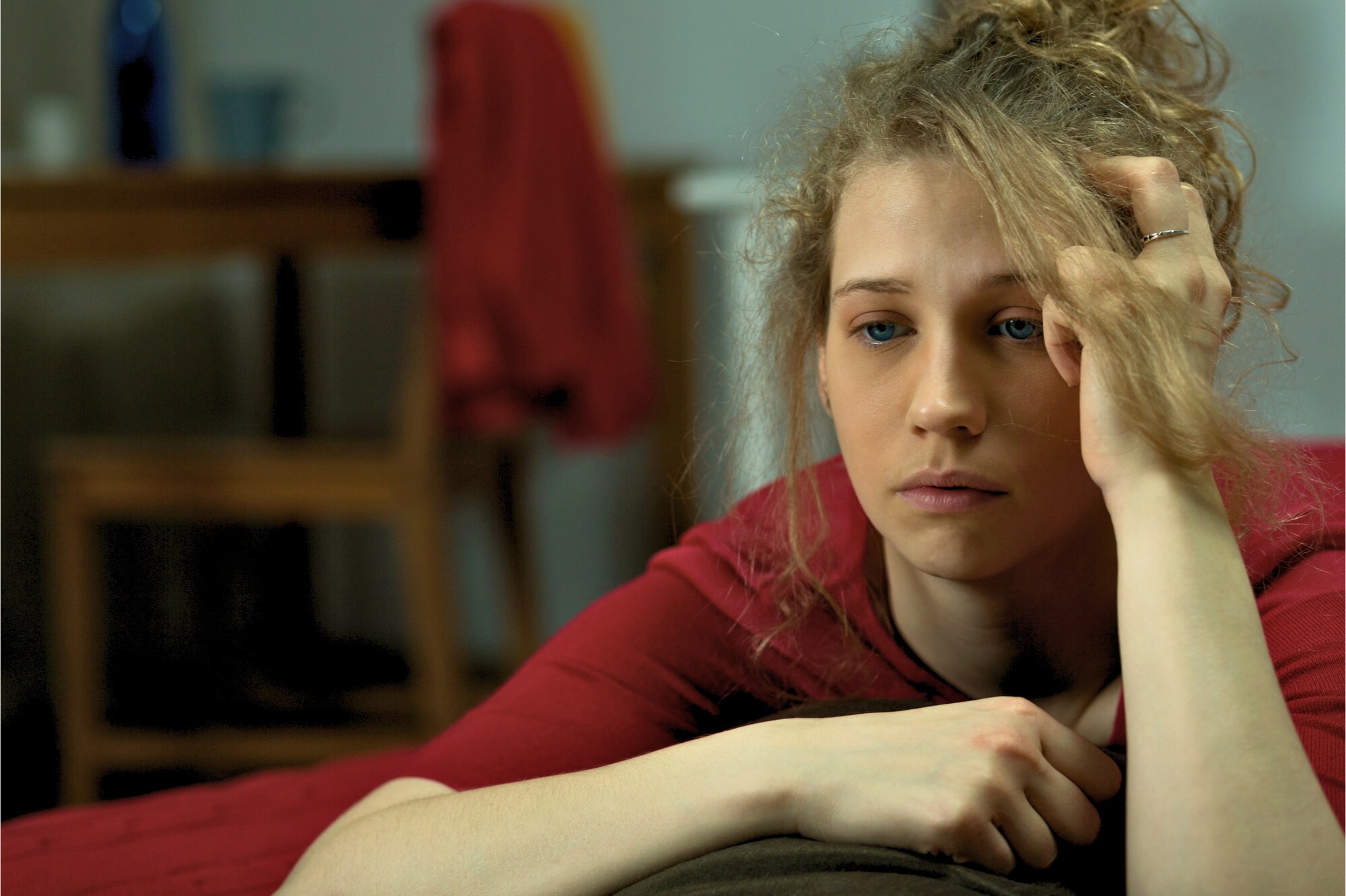 A young woman with blonde curly hair rests her head on her hand, looking contemplative and slightly sad. She wears a red top and is leaning on a surface with a blurred background that includes a chair and a red garment hanging.