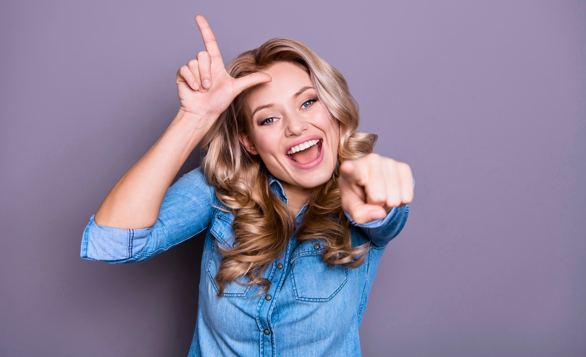 A cheerful woman with long, wavy blonde hair is smiling widely and making a playful gesture. She is forming an "L" shape with her right hand on her forehead while pointing forward with her left hand. She is wearing a light blue denim shirt. The background is purple.
.