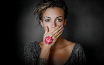 A woman covers her mouth with her hand, which has a red stop sign drawn on it. She looks directly at the camera against a dark, blurred background.