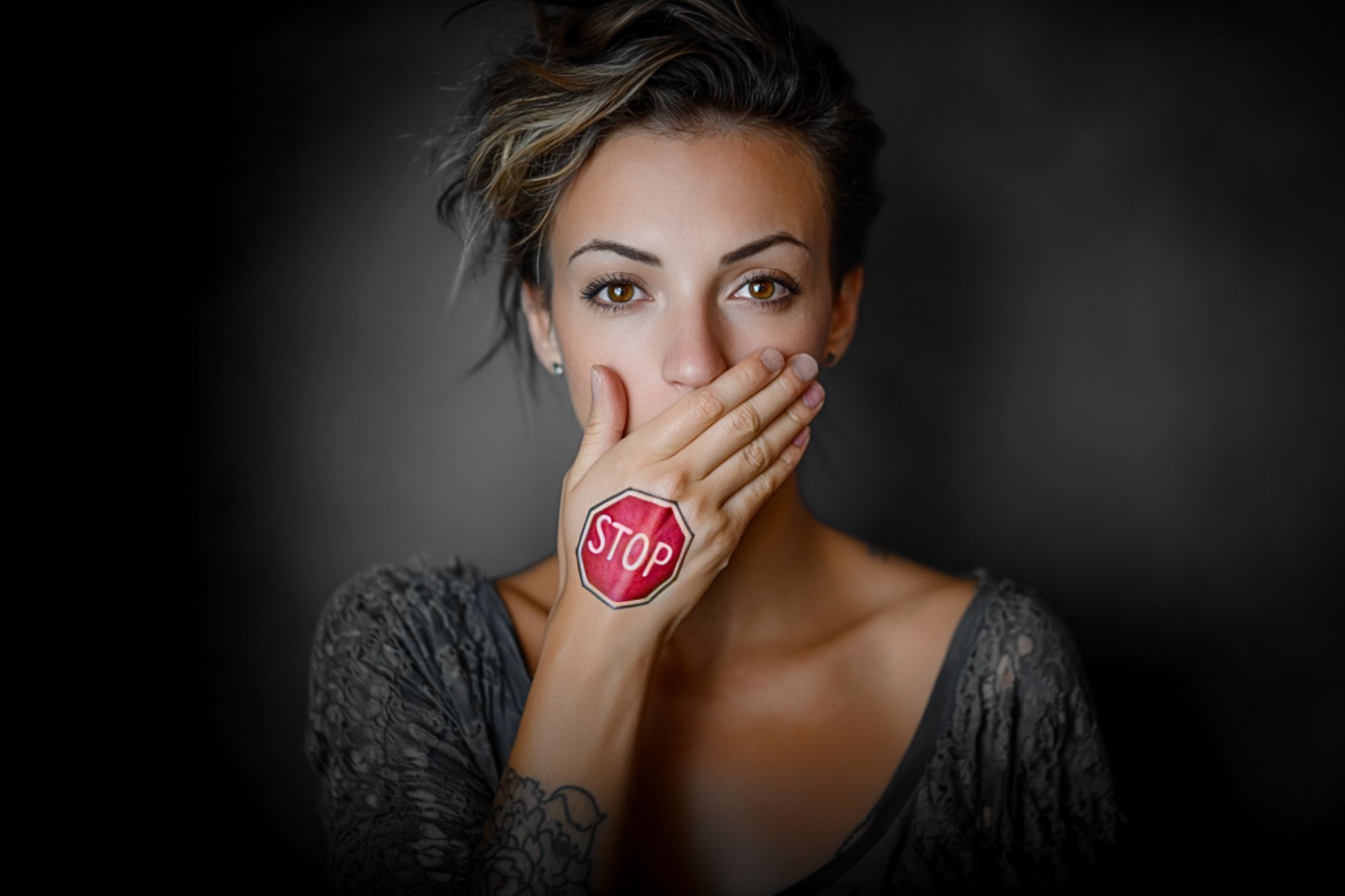A woman covers her mouth with her hand, which has a red stop sign drawn on it. She looks directly at the camera against a dark, blurred background.