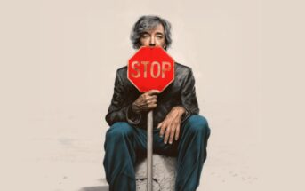 An older person with gray hair sits on a rock, holding a stop sign that covers their mouth. The background is plain and light-colored, drawing focus to the figure and the bright red sign.