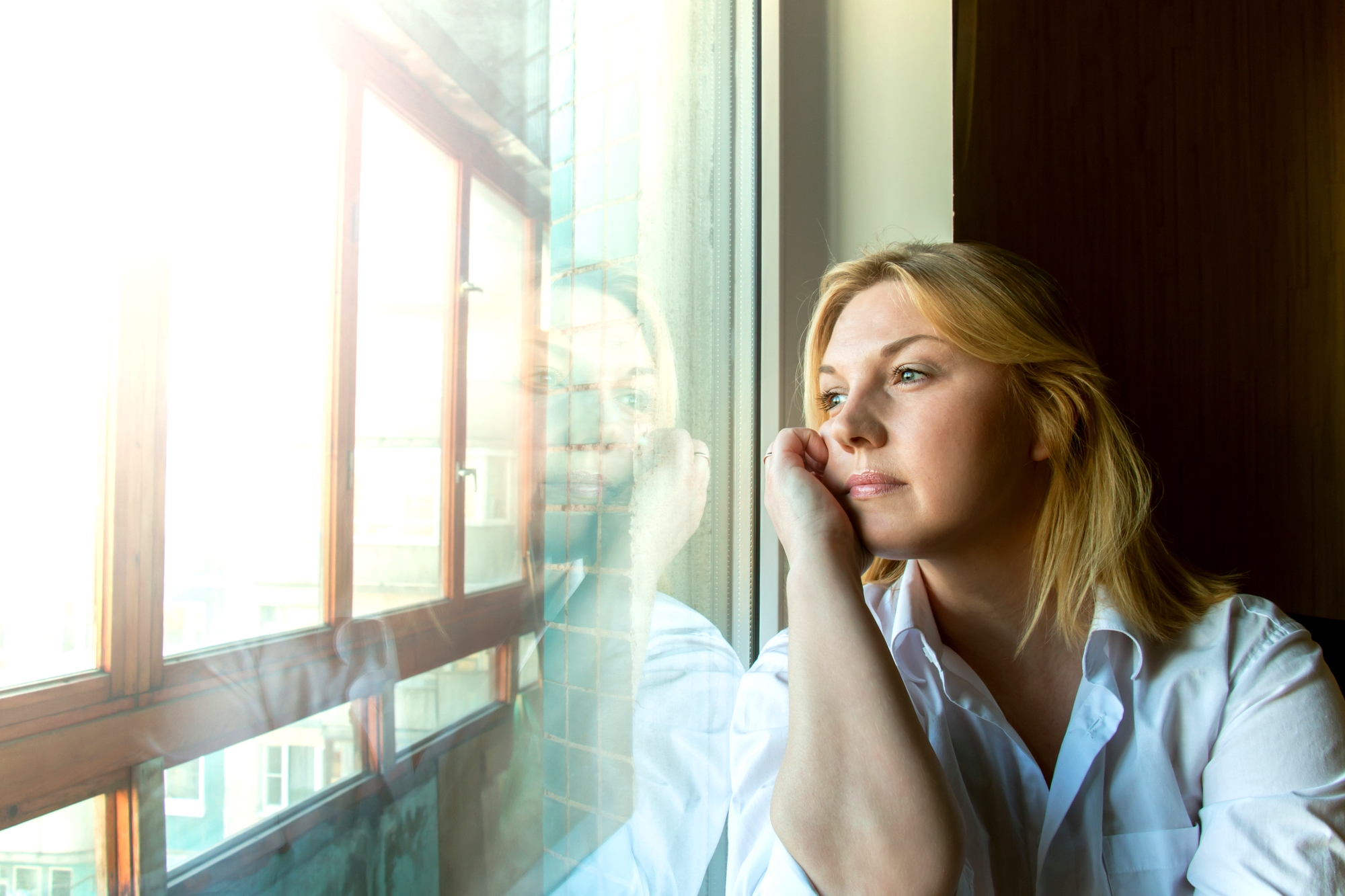 A woman with blonde hair, wearing a white shirt, rests her head on her hand and gazes thoughtfully out of a window. Sunlight streams through the window, creating a reflection of her face on the glass. Shadows suggest it's either early morning or late afternoon.