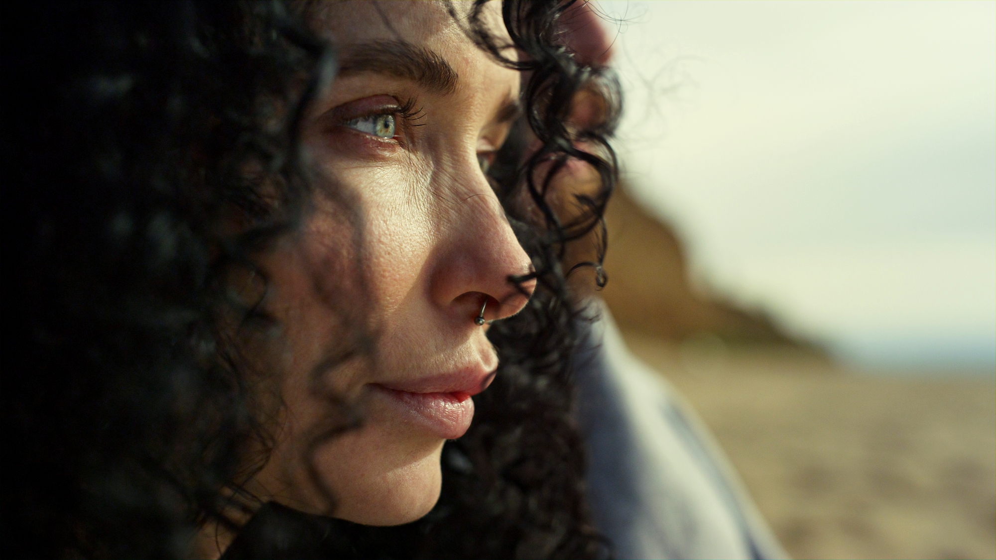 A close-up of a person with curly black hair, a septum piercing, and light-colored eyes, gazing into the distance. The background appears to be a sandy beach with blurred, sunlit scenery.