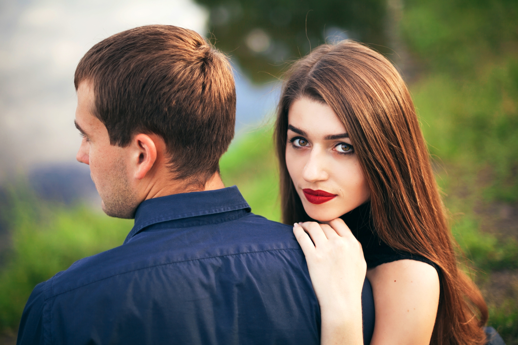 A woman with long brown hair and red lipstick rests her hand on the shoulder of a man with short brown hair, who is facing away from the camera. They are outdoors, with blurred green foliage in the background.