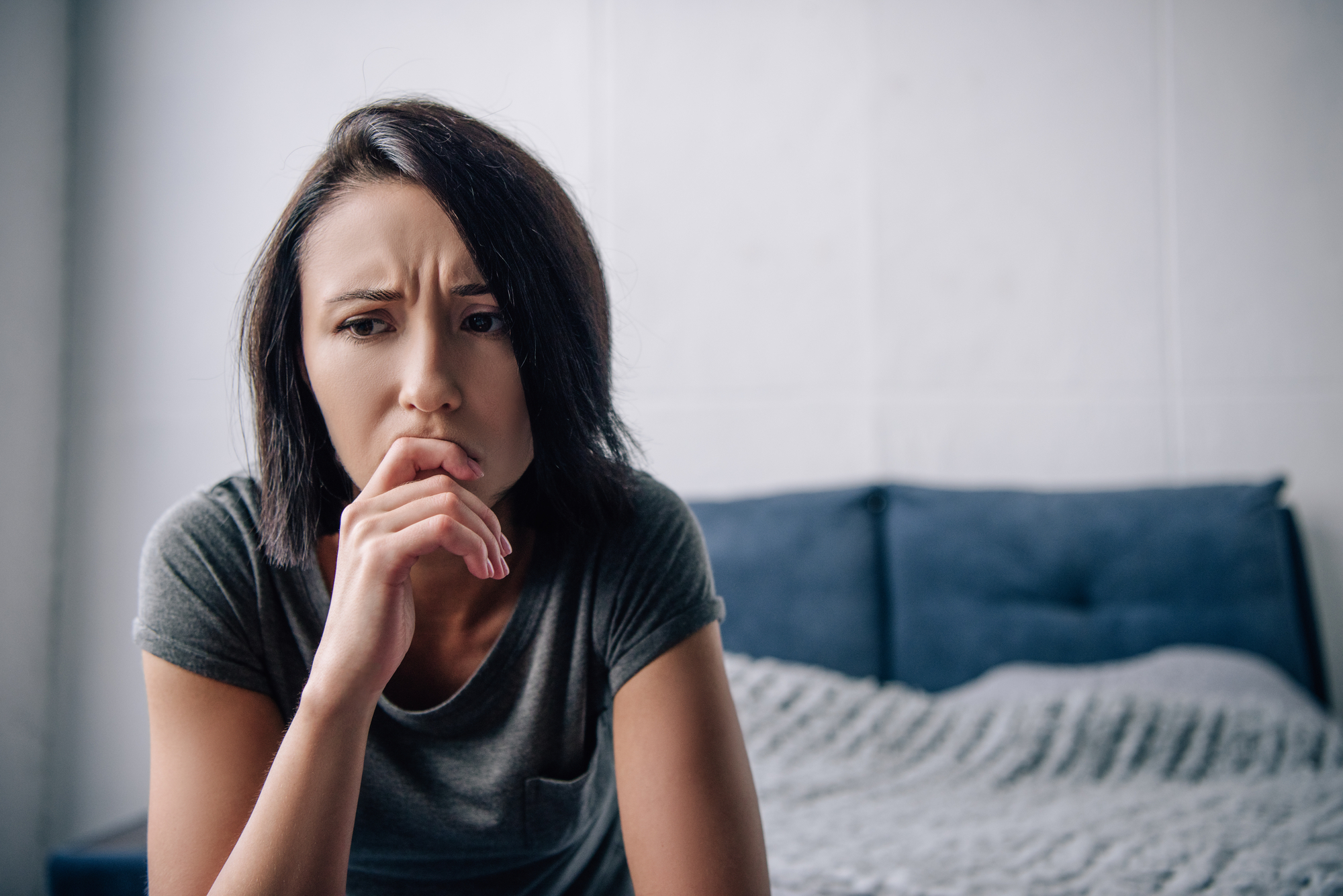 A woman with short dark hair looks pensive and worried, resting her chin on her hand. She is sitting on the edge of a bed with a blue headboard and a light-colored blanket. The background is softly lit with neutral tones.