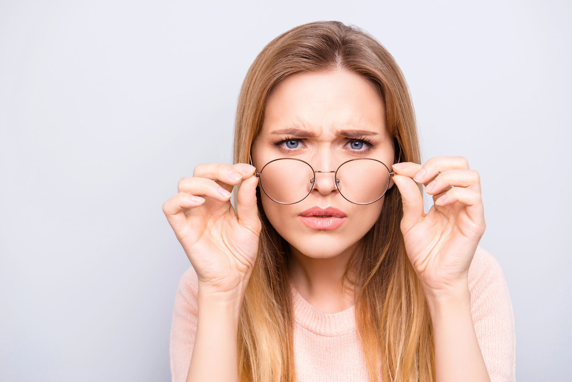 A woman with long blonde hair holds eyeglasses in front of her eyes and squints slightly, appearing confused or concentrating. Her facial expression is serious, and she wears a light pink sweater. The background is plain and light-colored.
