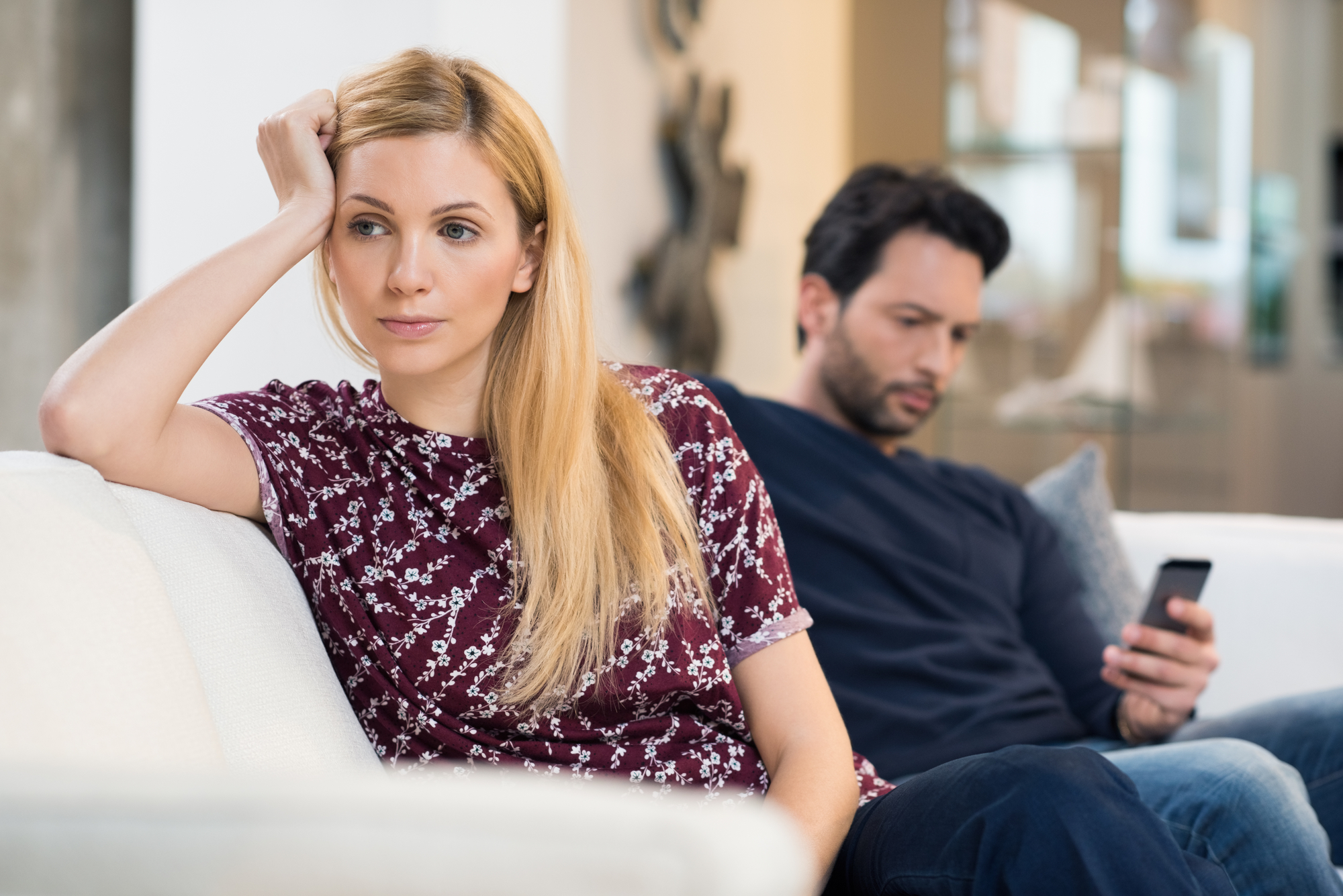 A woman with long blonde hair, wearing a maroon floral top, sits on a white sofa looking pensive, resting her head on her hand. A man with dark hair and a beard, wearing a navy blue shirt, sits behind her, focused on his phone.