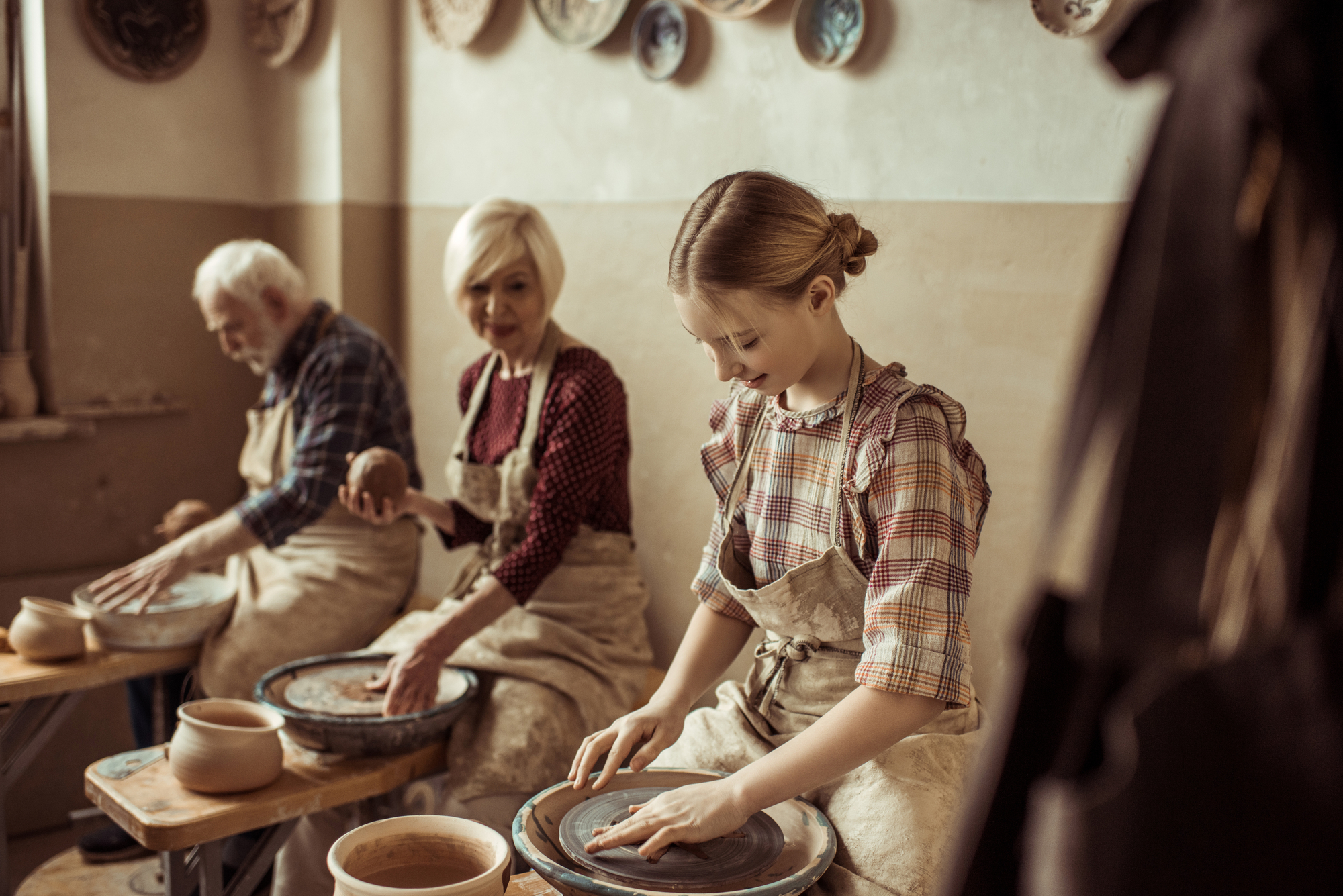 Three people are working on pottery wheels in a studio. A young girl in the foreground focuses on shaping clay, a woman in the center holds a ball of clay, and an elderly man in the background works on a clay piece. Various ceramic works adorn the walls.