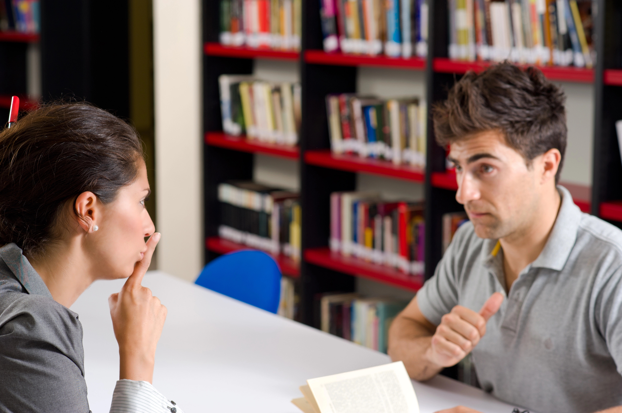 Two people are sitting across from each other at a table in a library. One person, facing the other, has a finger over their lips in a gesture for silence. The second person appears to be holding a book and gesturing with their thumb. Shelves filled with books are in the background.