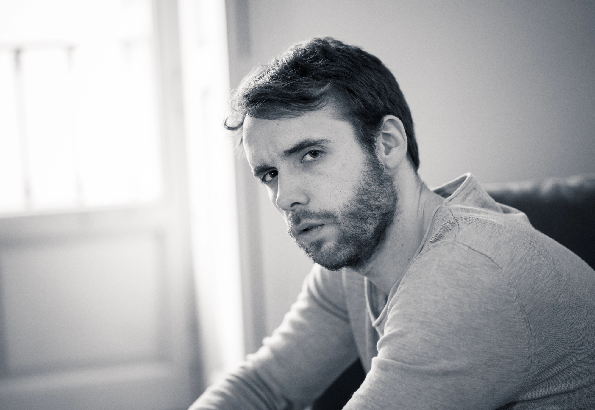 A black-and-white photo shows a man with short hair and a beard. He is wearing a light-colored shirt and is seated, leaning forward slightly, with a thoughtful expression on his face. The background is softly lit, with a window visible behind him.