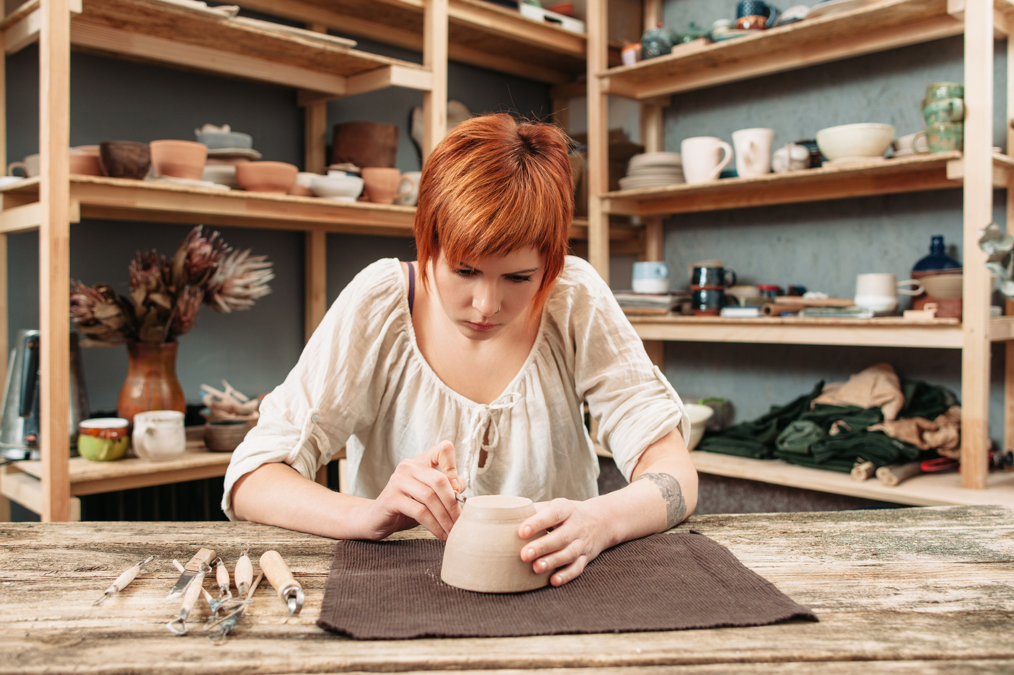 A person with short red hair is meticulously sculpting a clay pot on a table in a pottery studio. The individual is dressed in a white, long-sleeved shirt and surrounded by various pottery tools and shelves filled with pottery items in the background.