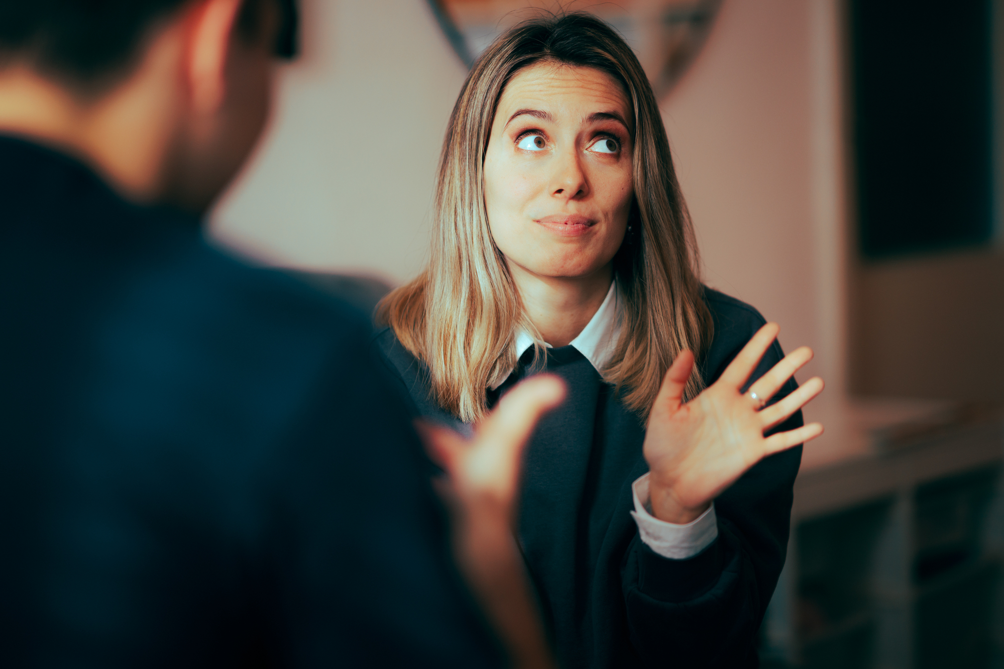 A woman with shoulder-length blond hair is gesturing with her hands up and a slightly exasperated expression on her face. She is sitting indoors and facing a person who is partially visible in the foreground. The setting appears to be casual.