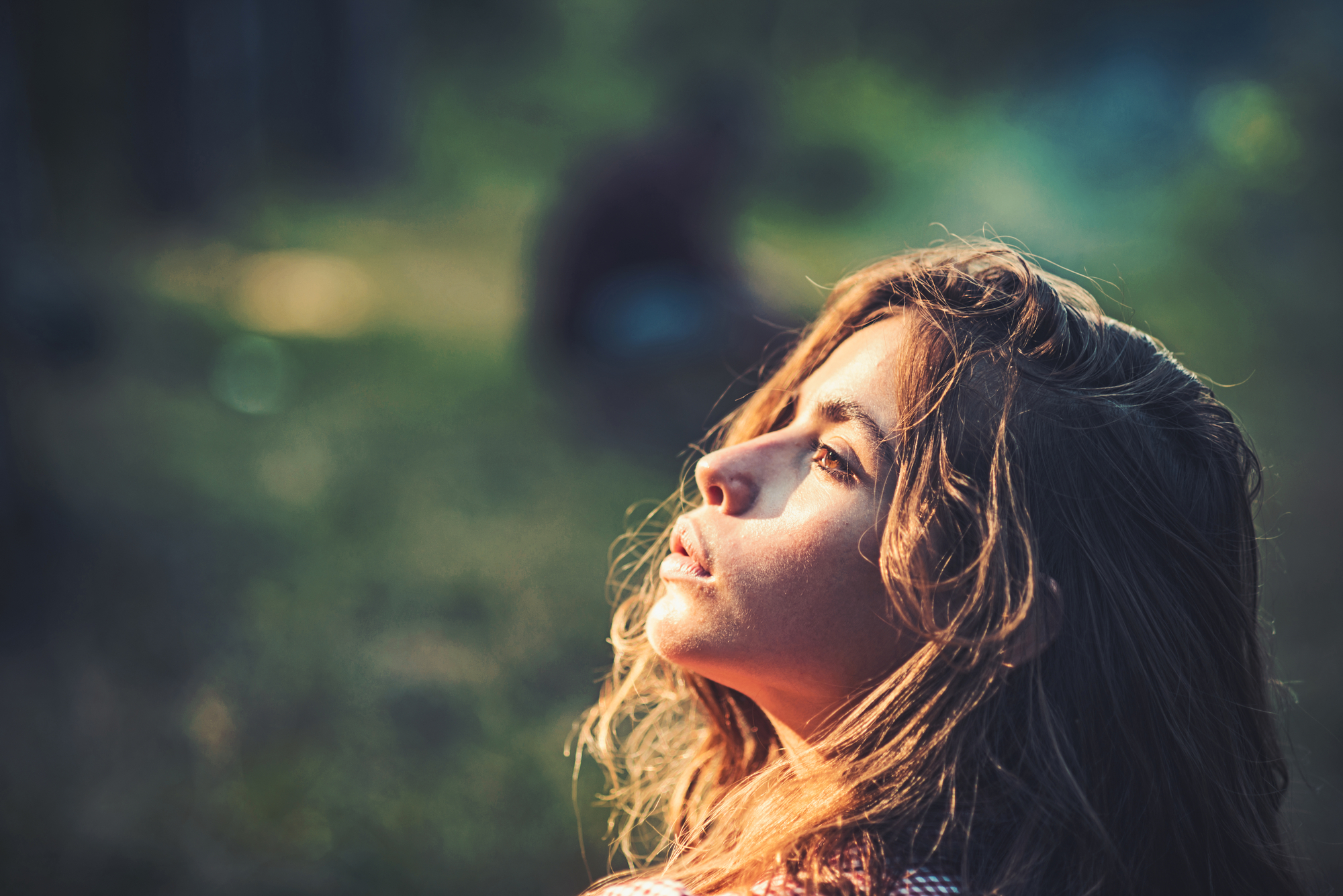 A young woman with wavy hair basks in the sunlight, eyes closed and face turned slightly upwards, appearing serene. The background is blurred, highlighting the contrasting play of light and shadow on her contemplative expression.