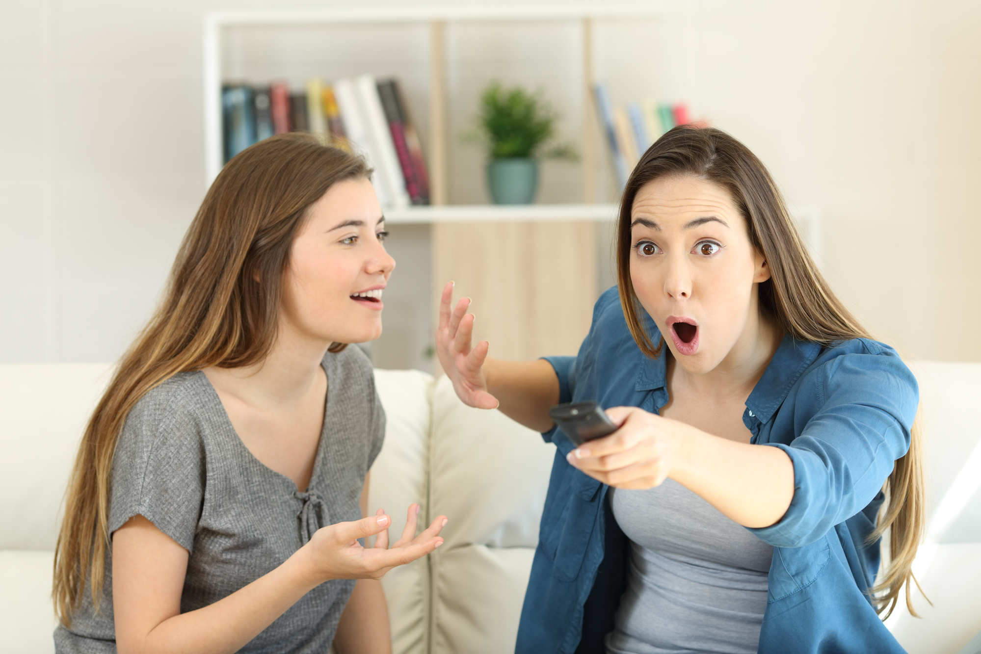 Two women sitting on a couch appear excited and surprised while watching something on TV. The woman on the left is smiling and gesturing with her hands, while the woman on the right is holding a remote control and has a wide-open mouth in amazement. Bookshelves and a plant are in the background.