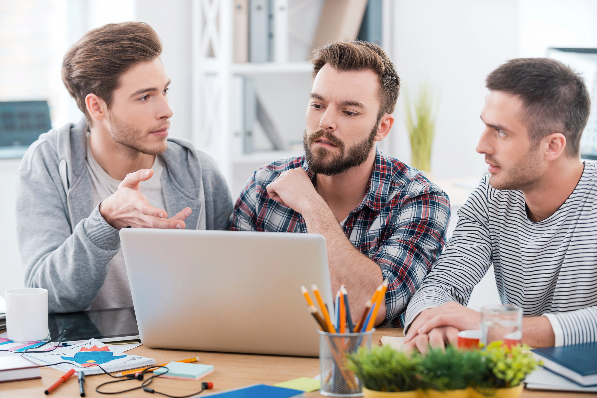 Three men are seated at a desk, engaged in a discussion. One man on the left is talking and gesturing with his hand. The man in the middle and the one on the right are listening attentively. A laptop, notebooks, and stationery are on the desk.