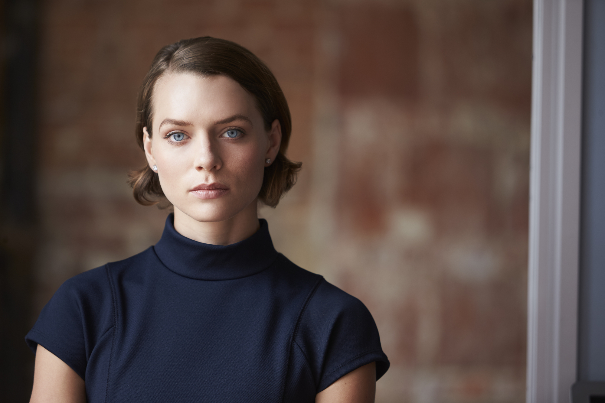 A woman with short brown hair, wearing a navy blue turtleneck top, stands indoors with a neutral expression. The background is softly blurred, comprising brown and beige tones.