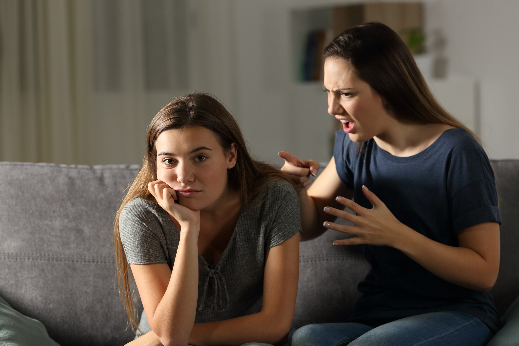 Two women are sitting on a couch. The woman on the left looks bored or uninterested, resting her chin on her hand. The woman on the right is animatedly talking or shouting at the other woman, with an intense facial expression and hand gestures.