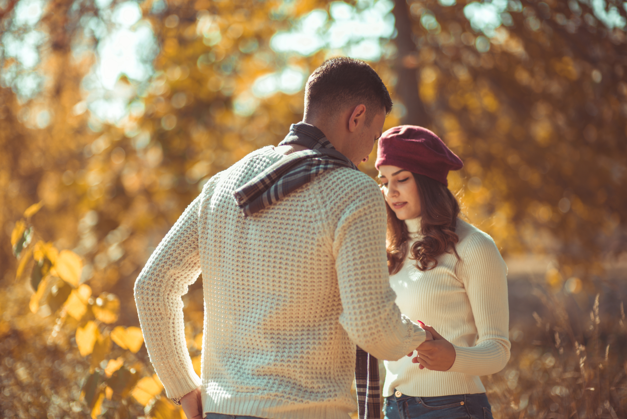 A couple stands outdoors among autumn foliage. The man, in a white sweater and scarf, gently holds the hand of the woman, who wears a white sweater and a red beret. They're engaged in a quiet, intimate moment with warm sunlight filtering through the trees.
