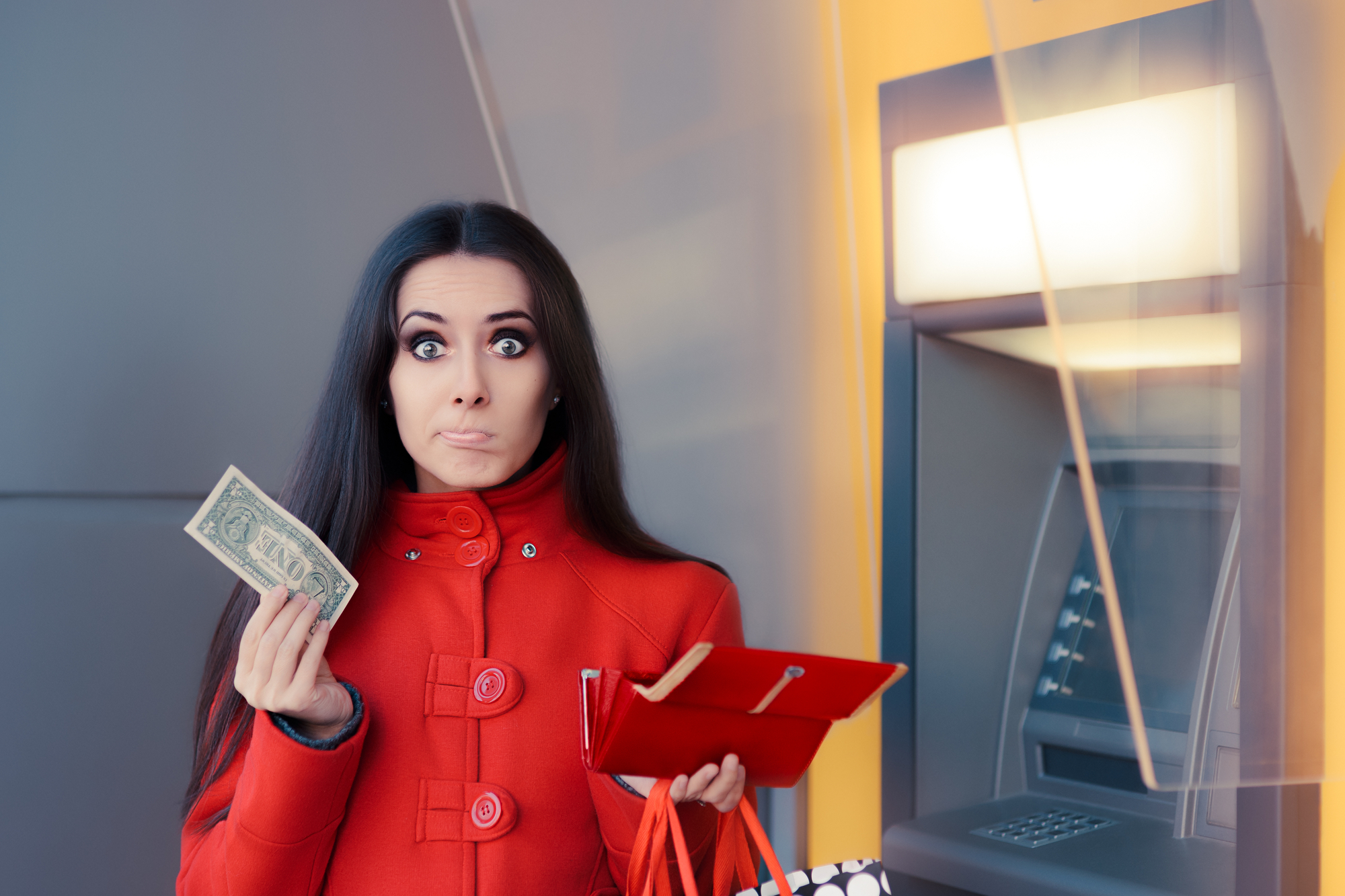 A woman with long dark hair, wearing a red coat, holds a single dollar bill in one hand and an open red wallet in the other. Her facial expression is of surprise or dismay. She stands next to an ATM, suggesting she might be experiencing a financial predicament.