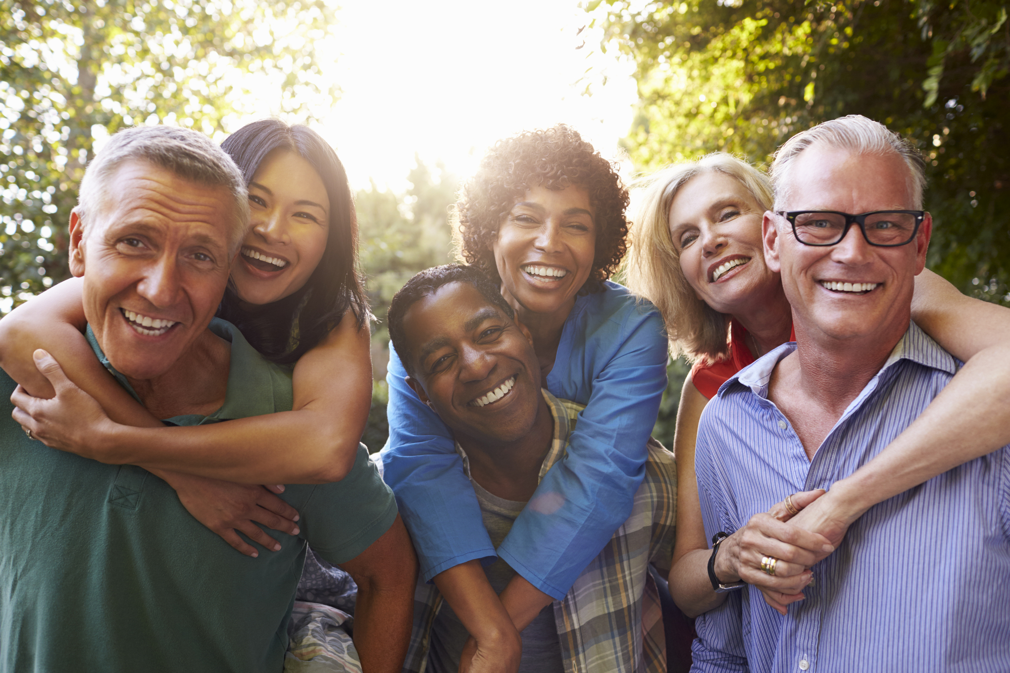 A diverse group of six adults smiling and posing cheerfully outdoors, with sunlight streaming through trees in the background. Two individuals in the front, one man, and one woman are being hugged from behind by two others. The atmosphere is lively and joyful.