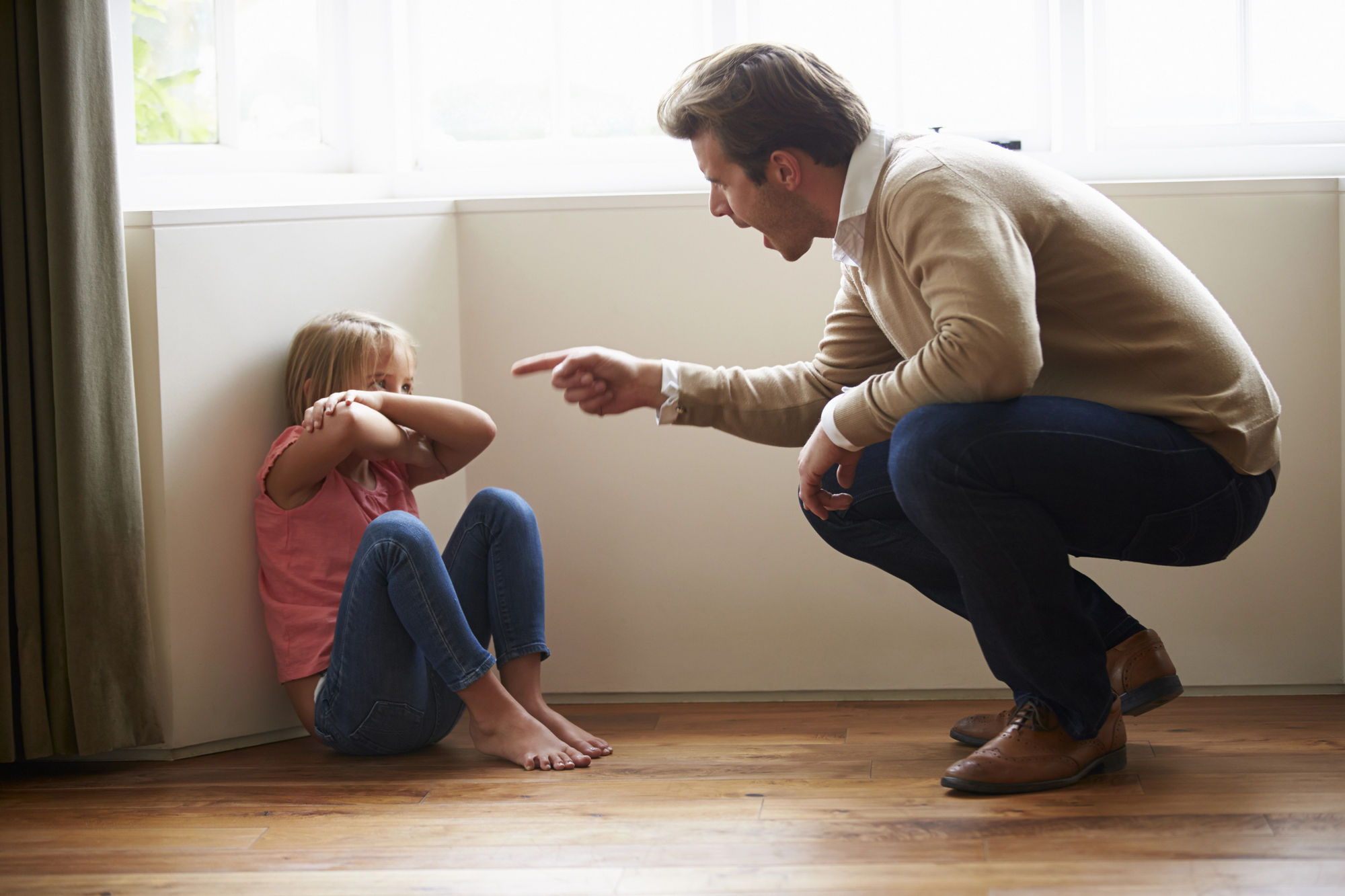 A man angrily points his finger at a young girl who is sitting on the floor against a wall, covering her ears with her hands. The man is squatting in front of her. They are in a room with wooden flooring and a large window in the background.