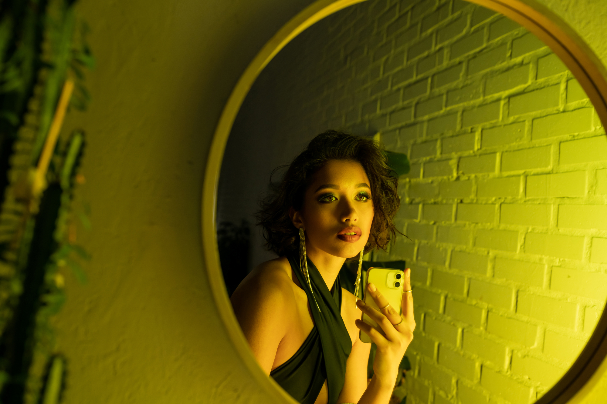 A woman with short curly hair is taking a selfie in a round mirror. She is wearing a green halter top and is surrounded by a yellow-green ambient light. There's a white brick wall in the background.