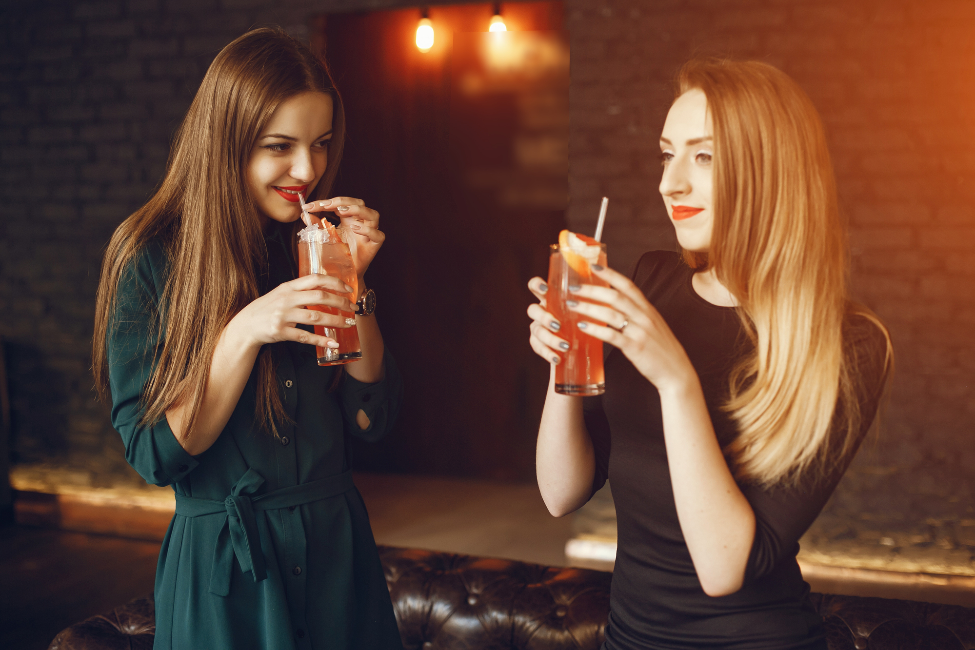 Two women are standing inside a dimly-lit space with exposed brick walls, each holding and sipping colorful cocktails. They are smiling and appear to be engaged in conversation, creating a warm and relaxed atmosphere.