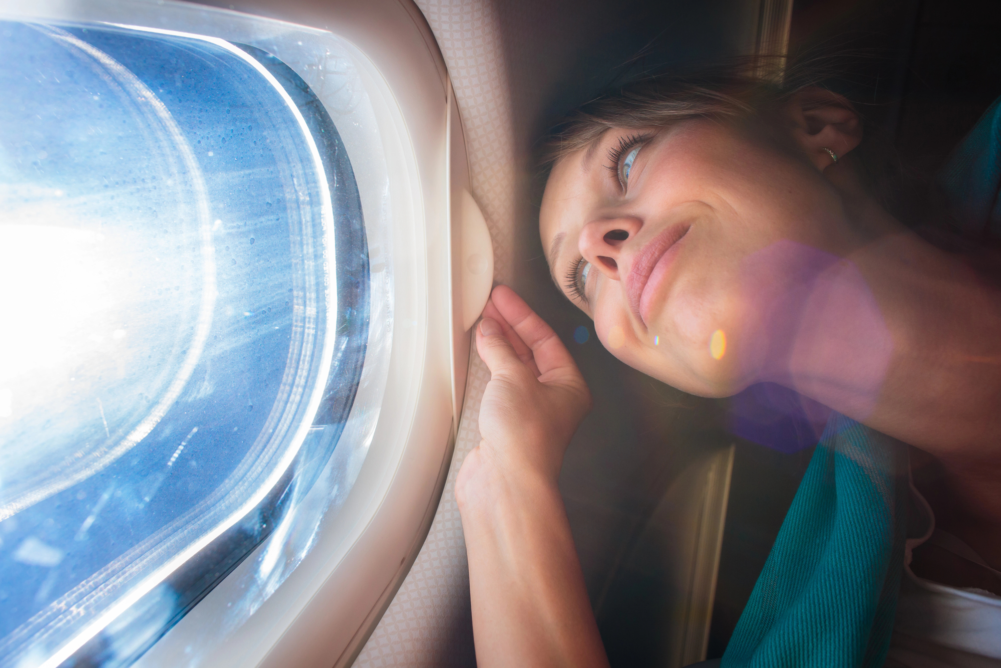 A woman leans close to an airplane window, gazing outside with a serene expression. Sunlight streams through the window, creating a lens flare effect. The window shows a view of the blue sky and white clouds.