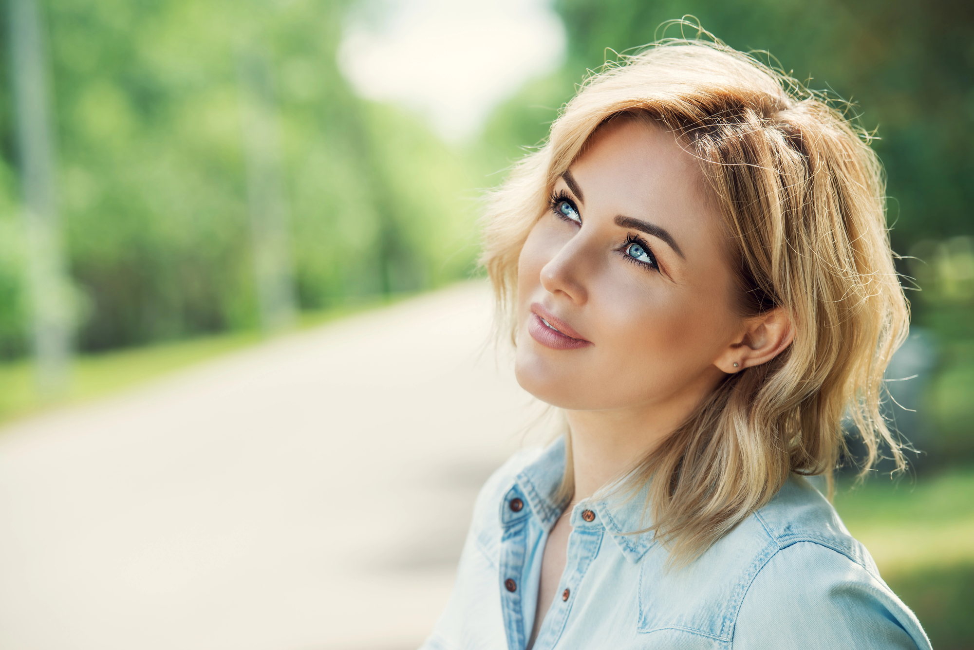 A woman with shoulder-length blonde hair and blue eyes dressed in a light blue denim shirt is standing outdoors. She is looking upwards with a soft smile. The background is blurred, showing a tree-lined walkway.