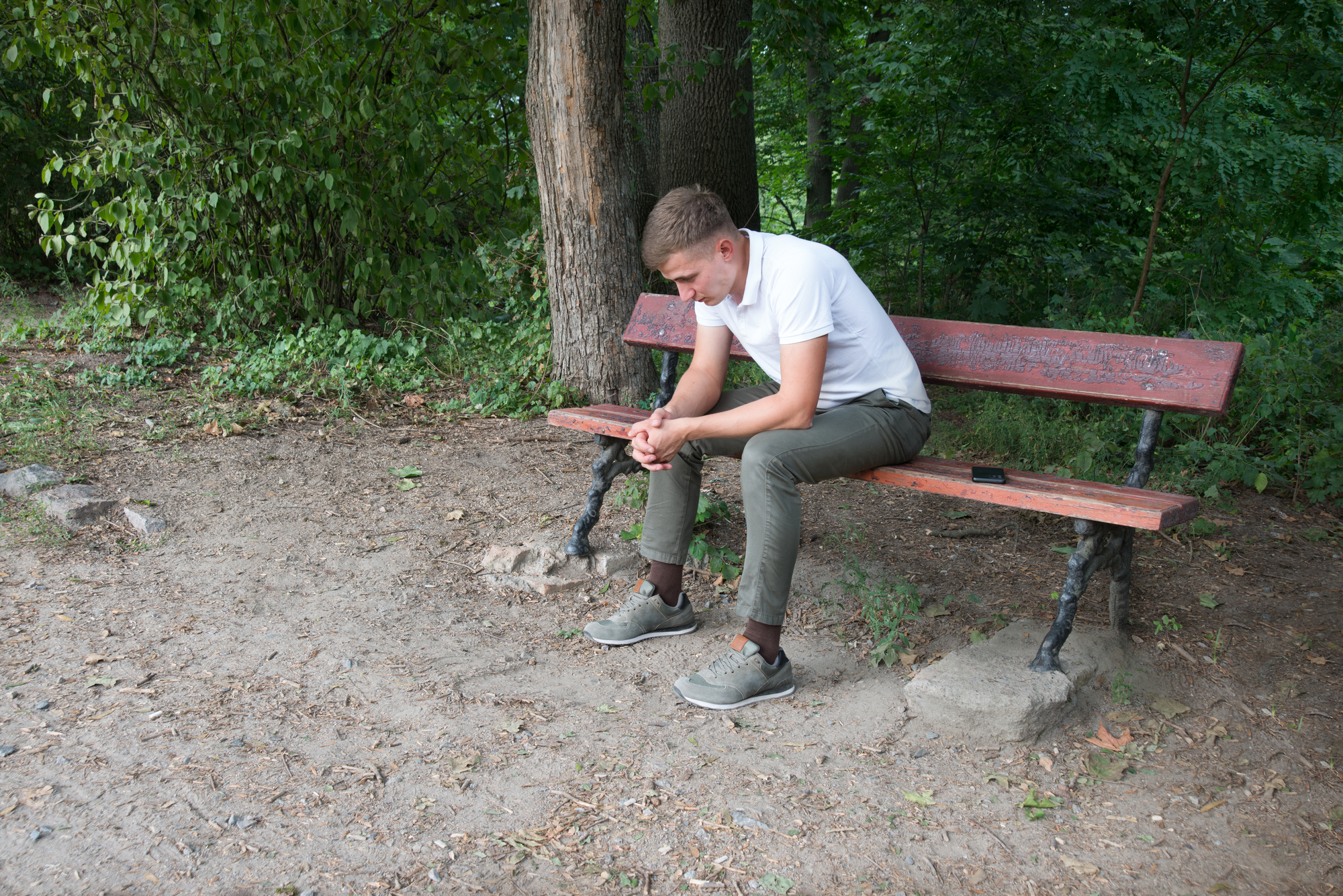 A young man with short hair sits on a red park bench in a forested area. He is wearing a white shirt and green pants, leaning forward with his elbows on his knees, appearing deep in thought. A phone lies on the bench beside him.