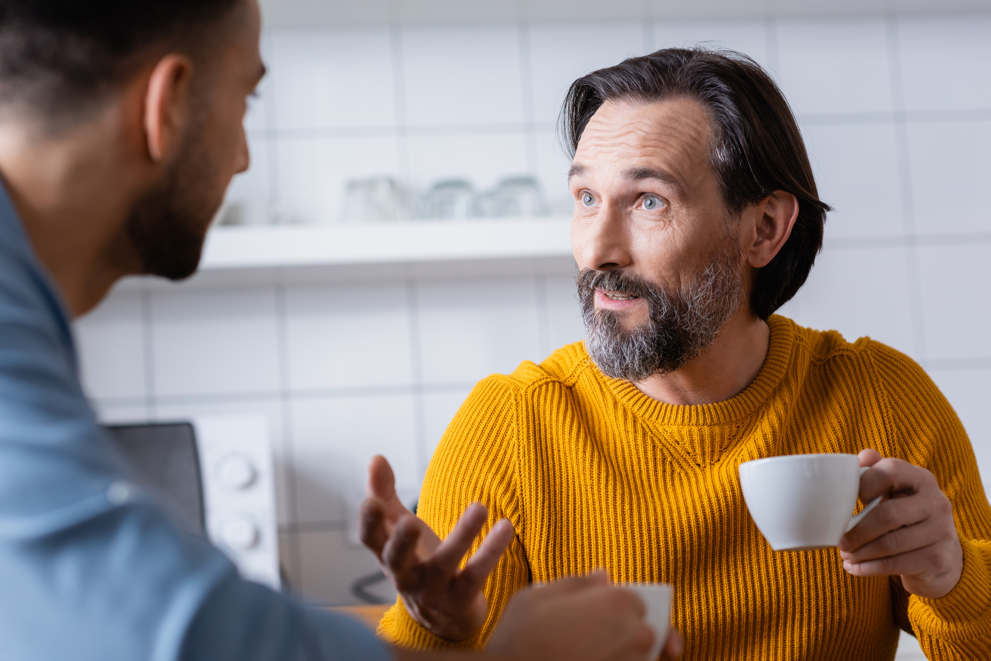Two men are having a conversation in a kitchen. The man on the right, wearing a mustard yellow sweater, is holding a white cup and appears to be speaking. The man on the left, facing away, is listening intently. The background shows white tiled walls and kitchenware.