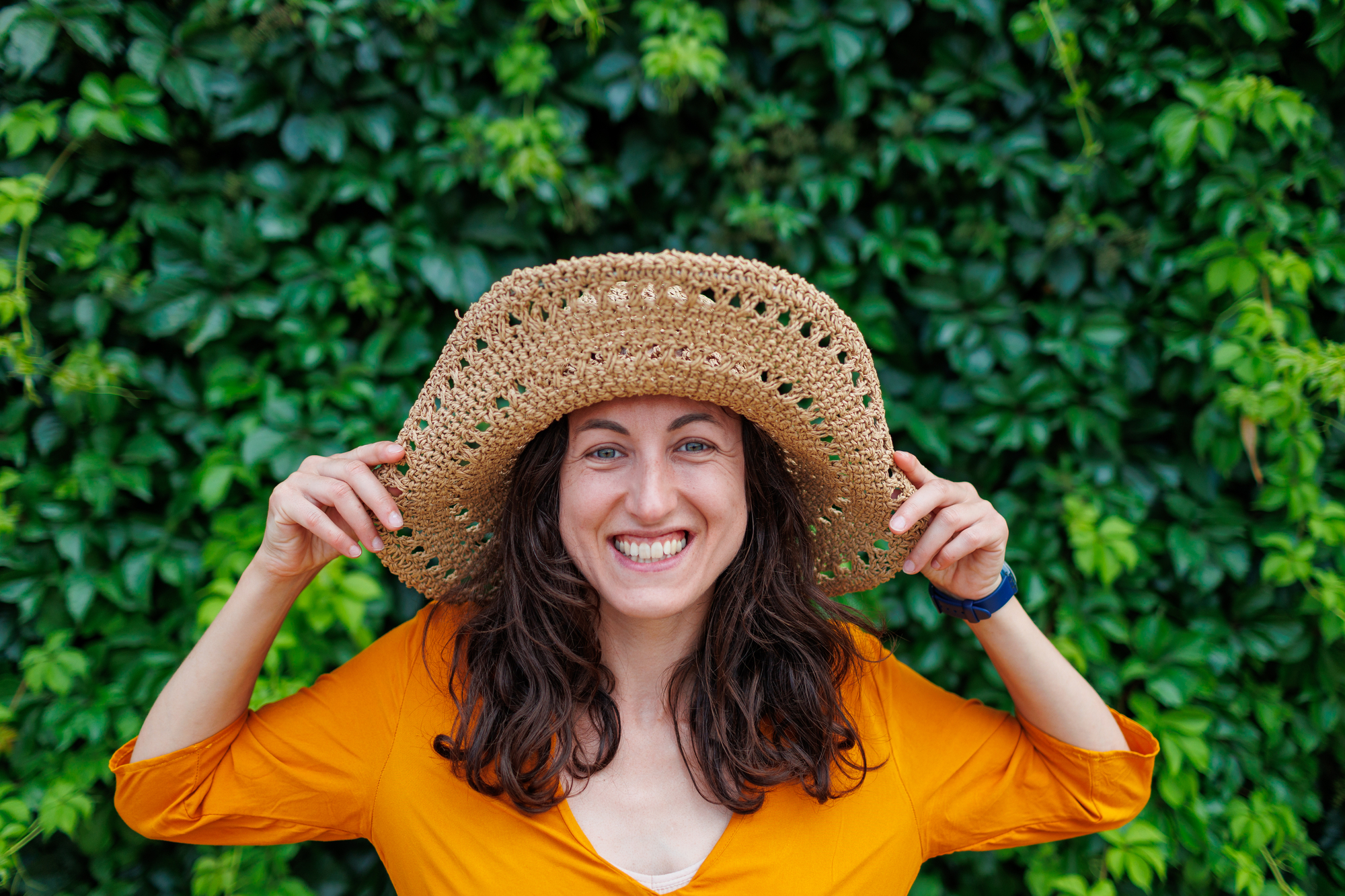 A smiling woman stands in front of a lush green background, wearing a large woven straw hat and an orange top. She has long, wavy brown hair and is holding the brim of the hat with both hands.