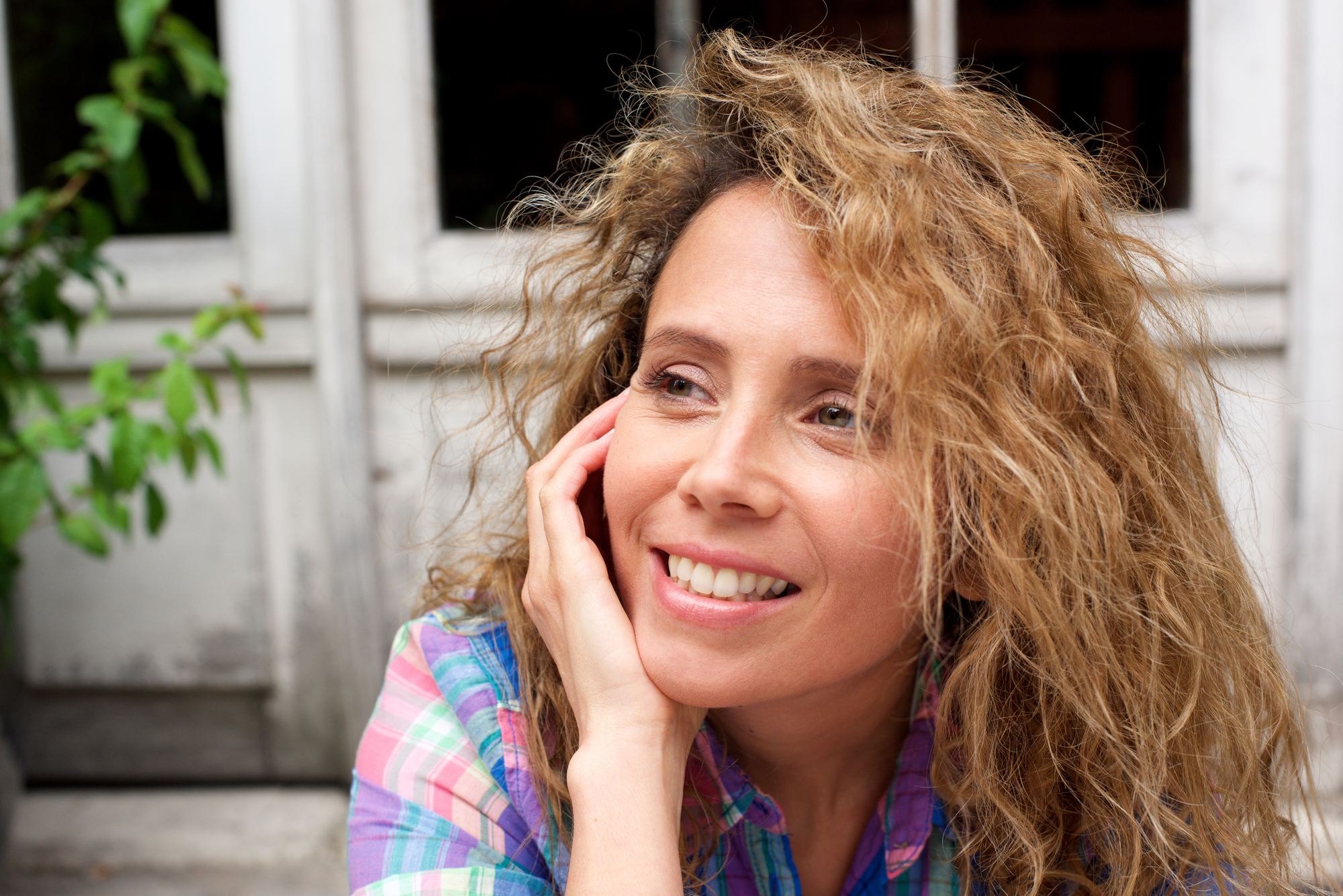 A woman with curly blonde hair, resting her chin on her hand, smiles while looking away from the camera. She is wearing a colorful plaid shirt and is sitting in front of a white window with a plant visible in the background.