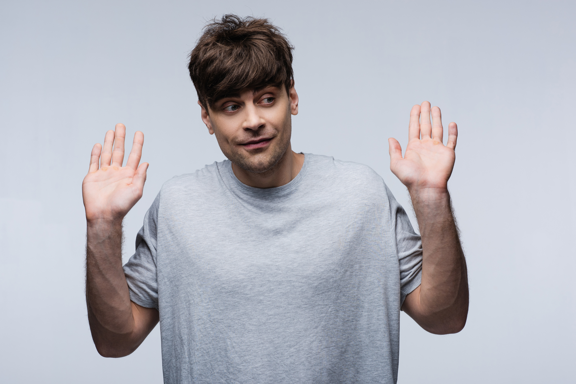 A person with short brown hair, wearing a light gray t-shirt, stands against a plain background. They are shrugging with both hands raised and palms facing outward, looking slightly to the side with a neutral expression.