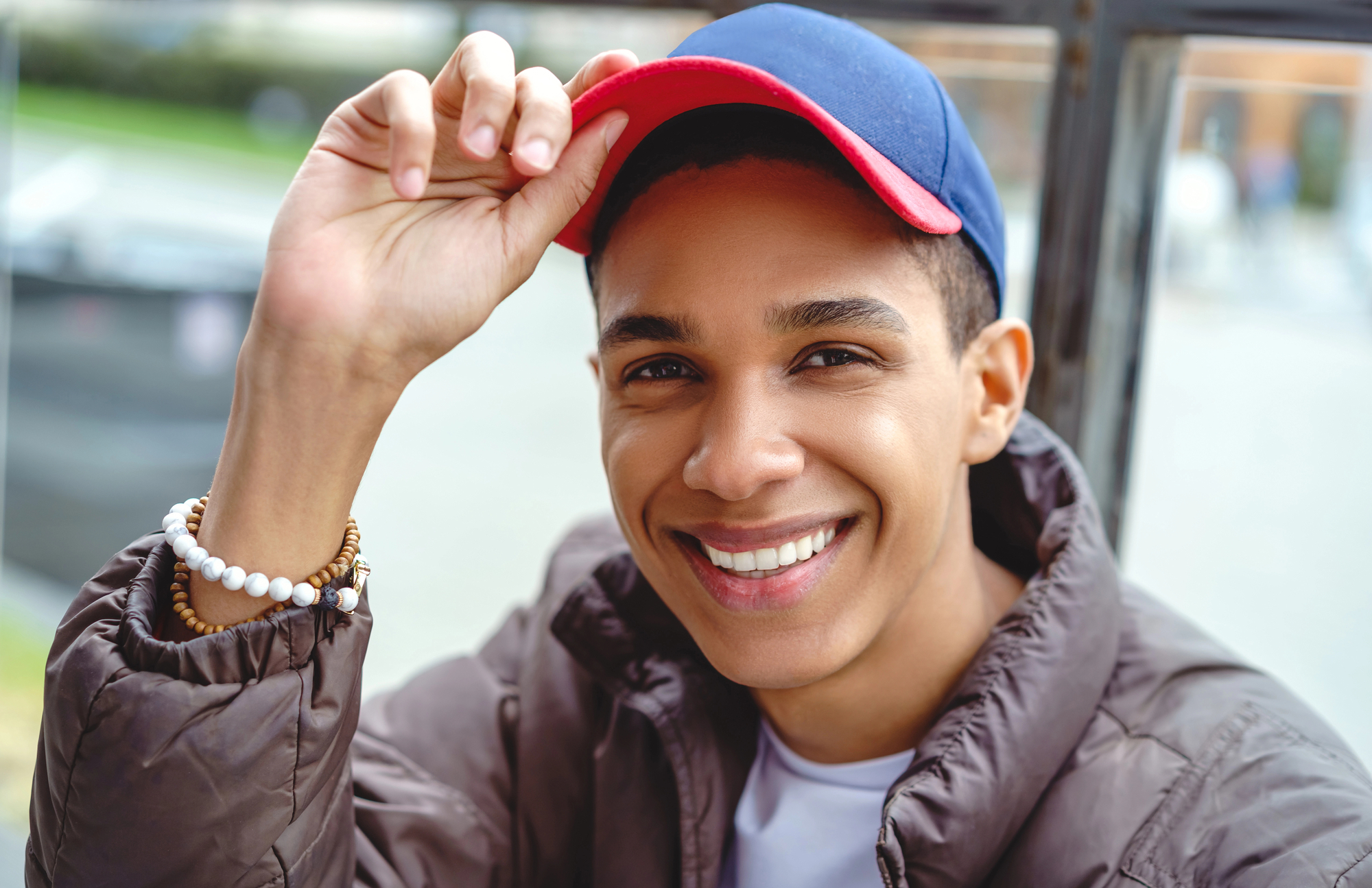 A smiling person wearing a blue and red baseball cap and a brown jacket is adjusting their cap with one hand. They have a beaded bracelet on their wrist and are sitting outdoors.