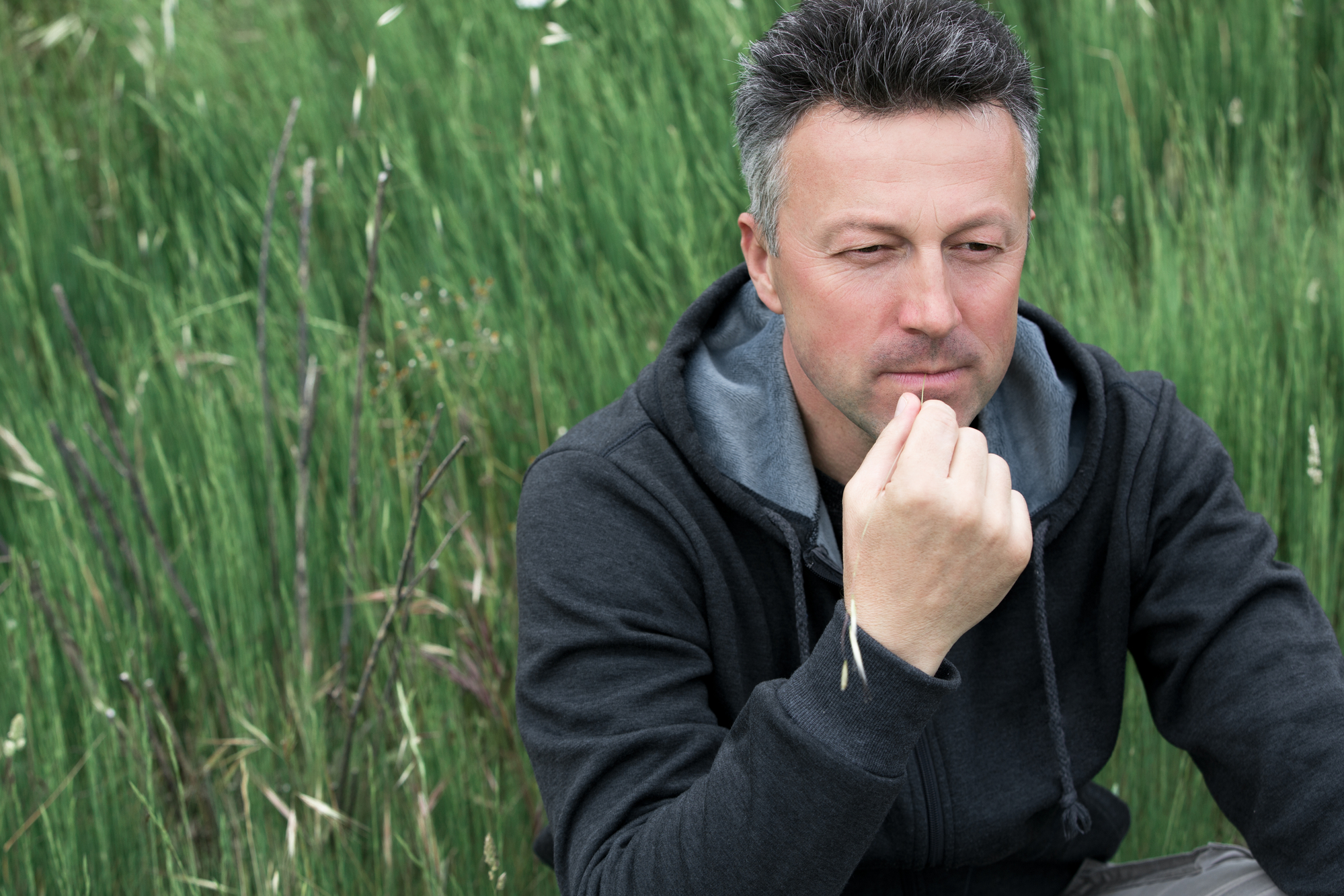 A man in a black hoodie sits pensively in a grassy field, holding his chin with one hand. He has short, dark-grey hair and a calm expression, looking slightly downward. The tall green grass surrounds him, creating a serene outdoor setting.